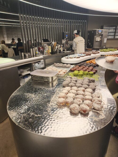 ステンレスを基調とした銀色の無機質な店内。曲線を描くカウンターと陳列されたドーナツ、スタッフの様子。