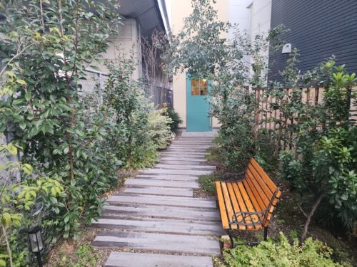 緑の植栽に囲まれた小道の奥にある、鮮やかな青い扉が特徴的なソラアオの店舗入り口。